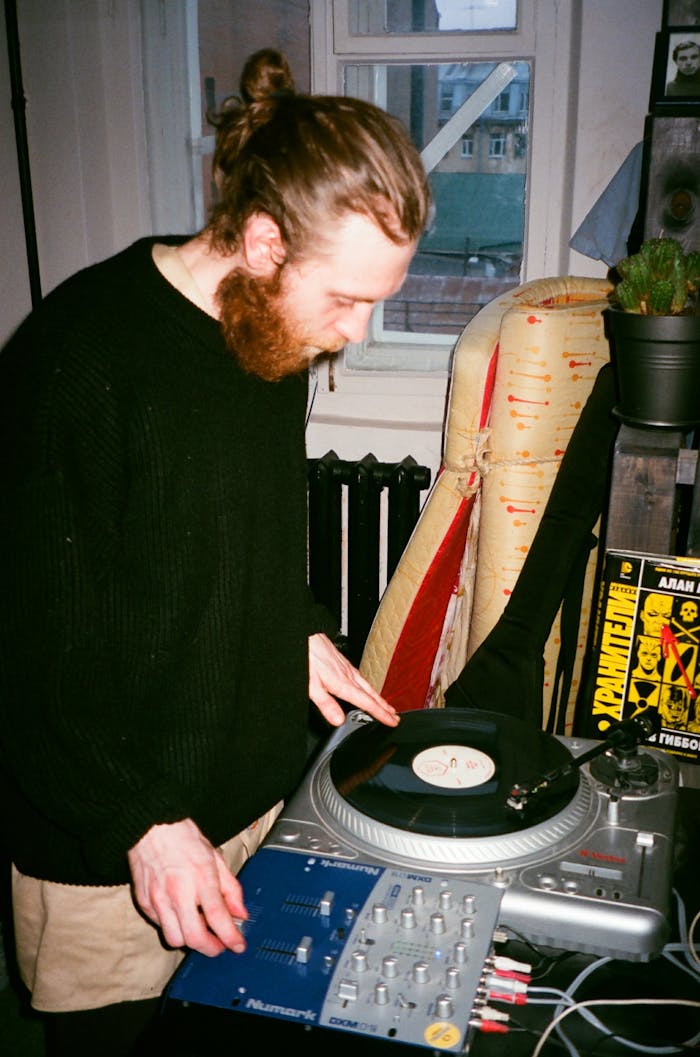 gallery-04 Man with beard and bun spinning vinyl on a turntable indoors in a cozy urban setting.